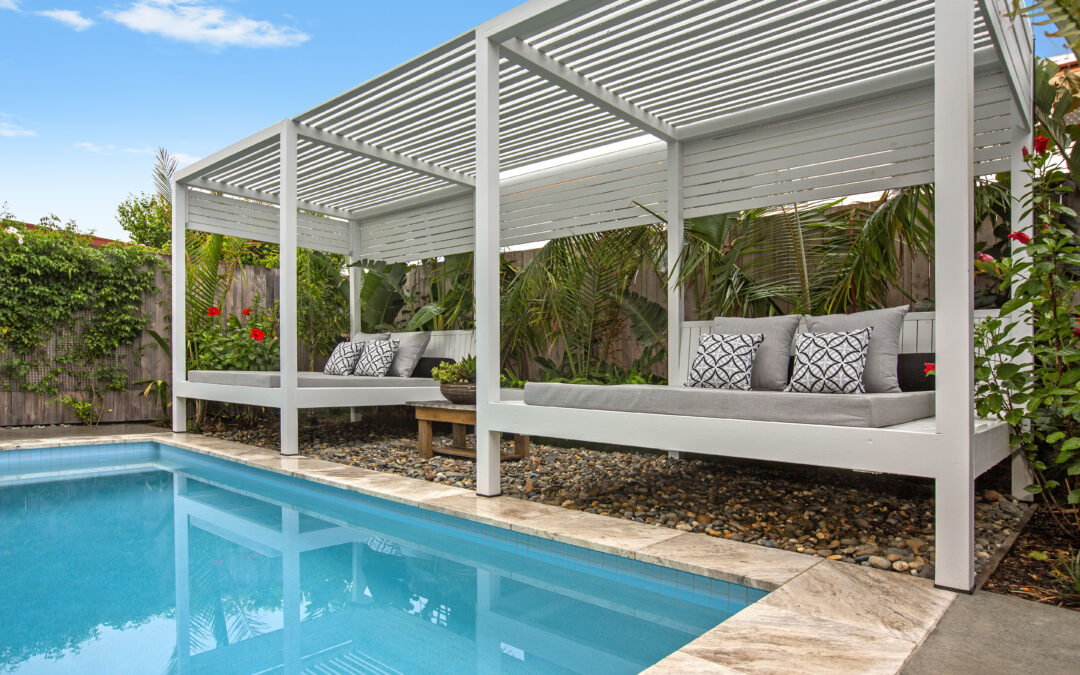 Modern poolside lounge with white pergolas, gray cushions, and tropical plants beside a blue swimming pool.