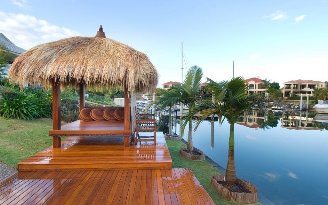 A wooden deck with a thatched-roof gazebo and built-in seating overlooks a calm waterfront canal lined with palm trees, boats, and modern houses under a clear blue sky.
