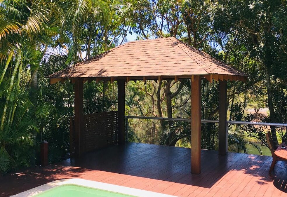 A wooden gazebo with a shingled roof stands on a raised deck beside a small pool, surrounded by lush tropical trees and greenery in bright daylight.