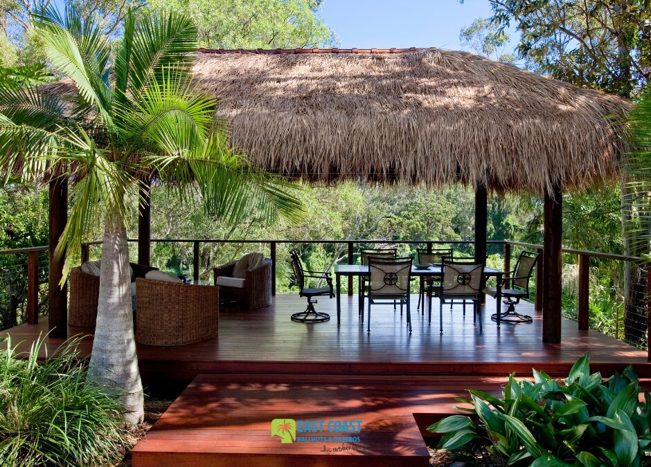 Tropical Bali hut with a natural thatched roof on a raised timber deck, featuring an outdoor dining set and lush garden surroundings.
