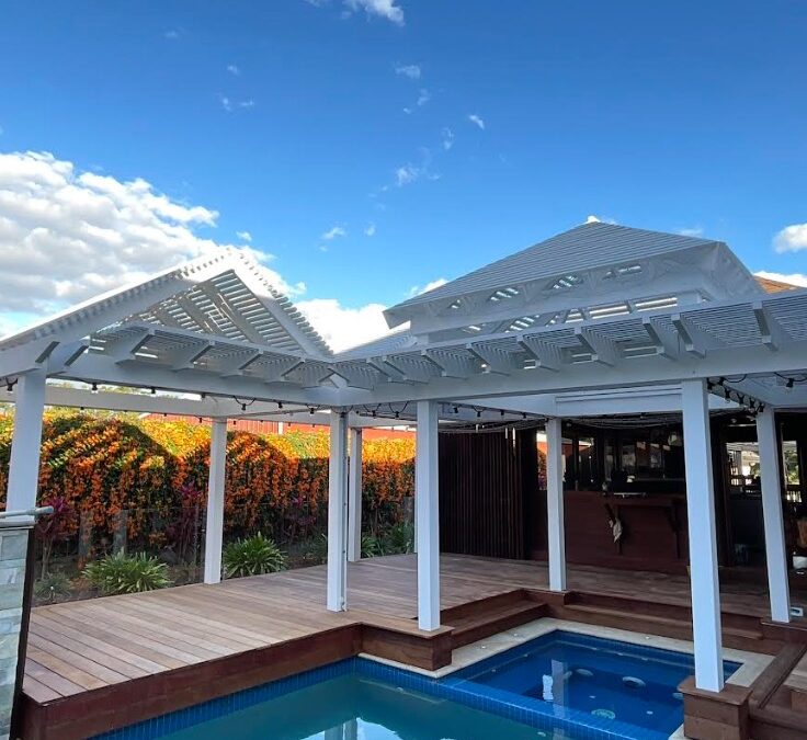 White pergola with louvred roof panels beside a swimming pool on a modern timber deck under a bright blue sky.